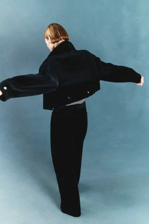 Model seen from behind with arms outstretched wearing a cropped black jacket and black trousers against a muted blue studio backdrop.