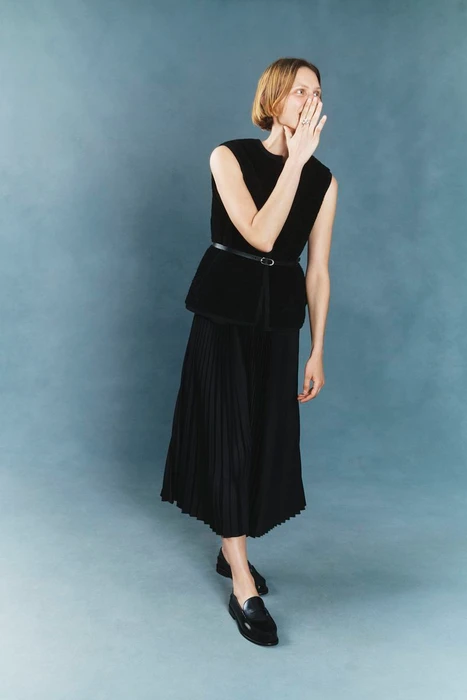 Model Indre Aleksiuk wearing a sleeveless black belted top, pleated midi skirt, and black loafers against a blue backdrop.