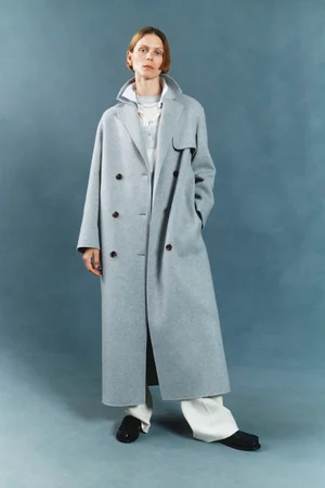 Model wearing a long grey double-breasted coat and white trousers standing against a textured blue backdrop