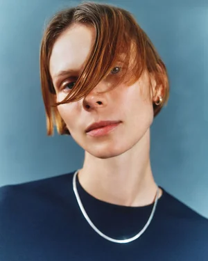 Close-up portrait of model Indre Aleksiuk with short auburn hair and a silver chain necklace wearing a navy top against a blue backdrop.