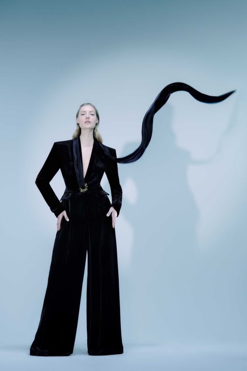 Elegant woman in a black velvet pantsuit poses against a light blue backdrop, accented by a flowing black ribbon detail.