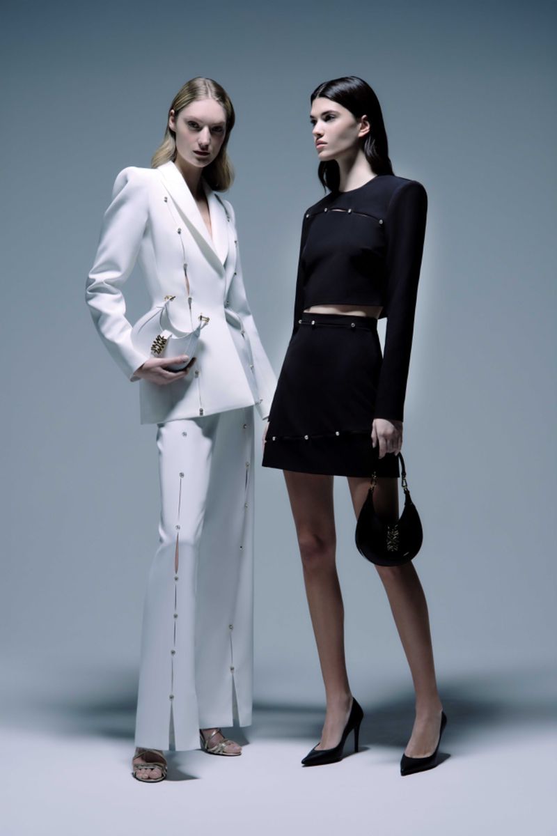 Two fashion models pose in a studio wearing matching black-and-white outfits, heels, and handbags. Fashion editorial shot.
