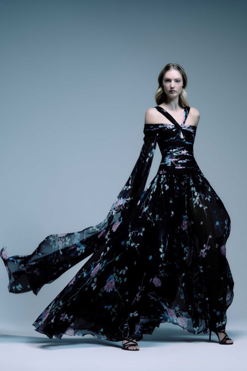 Woman in a flowing black gown with floral print posing against a light blue background in a studio for a fashion editorial shot.