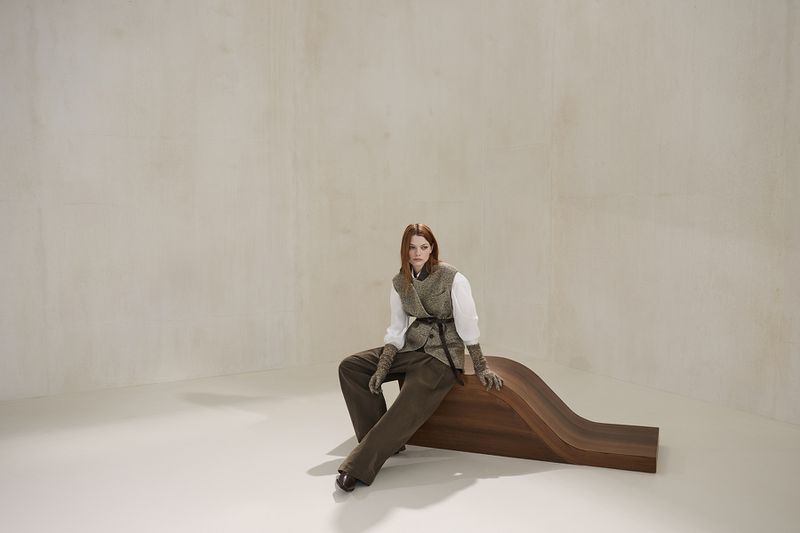 A fashionable woman poses in vest and trousers on a wooden bench in a photo studio with soft lighting and a minimalist backdrop.