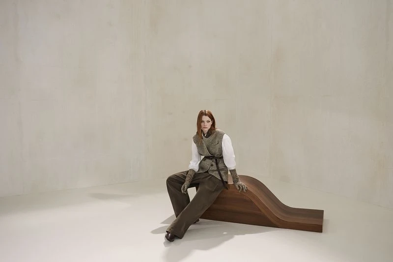 A fashionable woman poses in vest and trousers on a wooden bench in a photo studio with soft lighting and a minimalist backdrop.