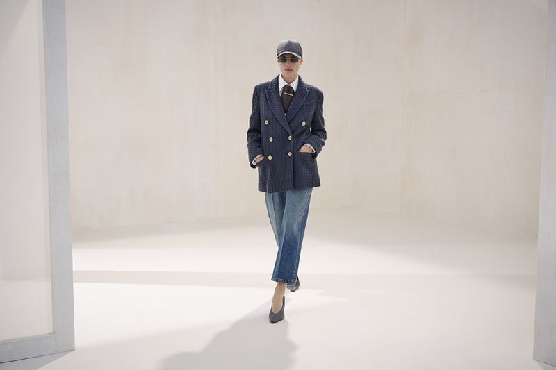 Model wearing a navy double-breasted blazer, cropped denim jeans, cap, sunglasses, and heels walking across a pale studio set.