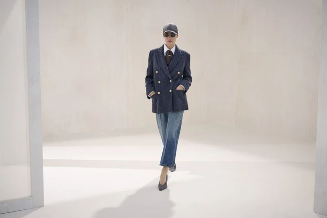 Model wearing a navy double-breasted blazer, cropped denim jeans, cap, sunglasses, and heels walking across a pale studio set.