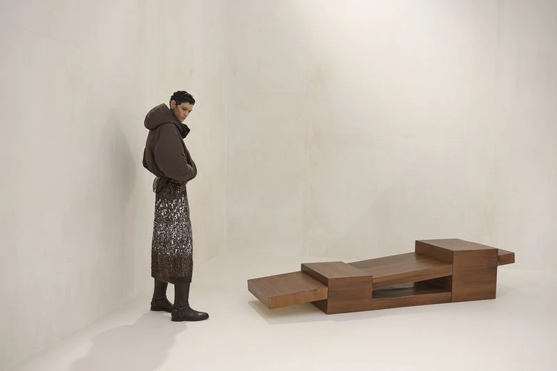 Fashionable adult in brown puffer jacket and patterned skirt poses next to a modern wood block bench in a bright photo studio.