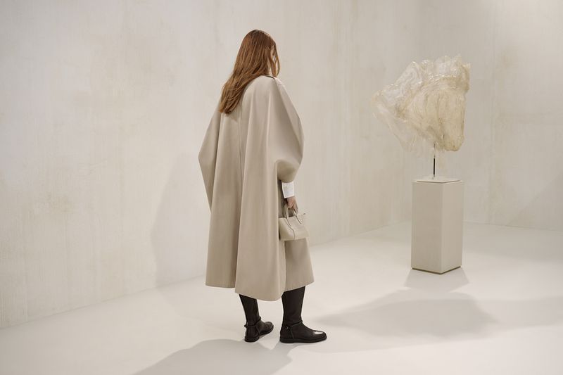 Woman in beige cape-coat and handbag standing in bright studio space with abstract art on a pedestal, facing away from the camera.