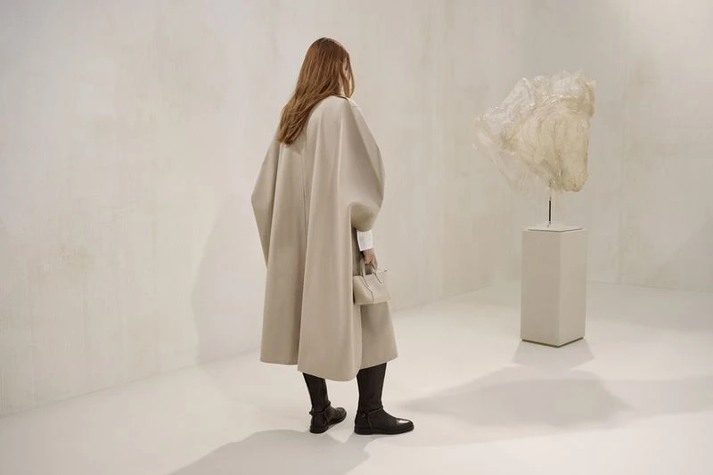 Woman in beige cape-coat and handbag standing in bright studio space with abstract art on a pedestal, facing away from the camera.