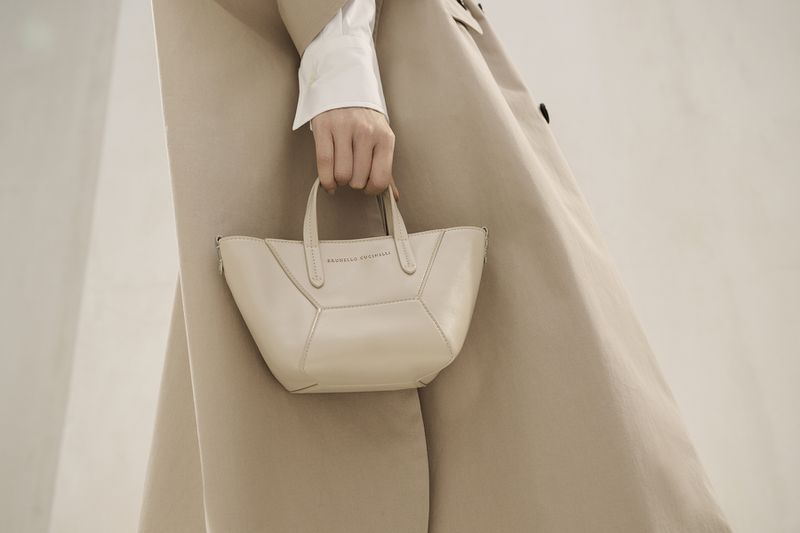 Cream-colored leather handbag held by a woman wearing a trench coat and white top, presented against a soft, neutral backdrop.