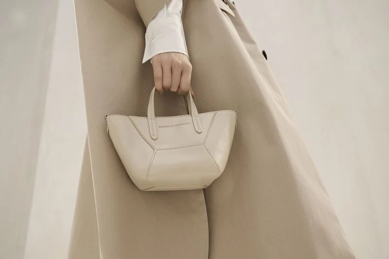 Cream-colored leather handbag held by a woman wearing a trench coat and white top, presented against a soft, neutral backdrop.