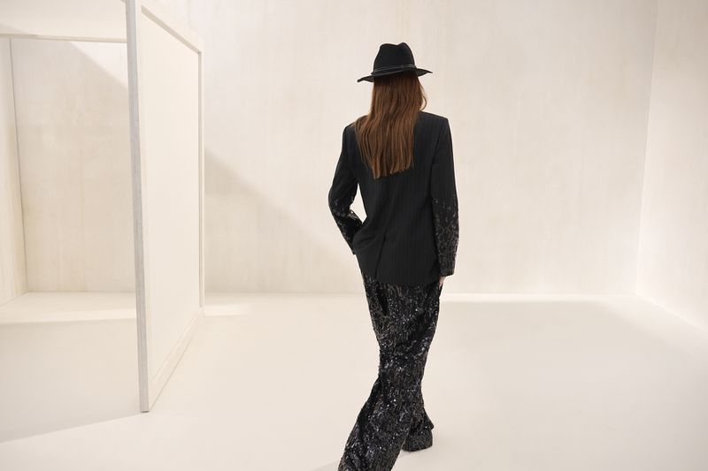 Back view of a fashionable woman wearing a black blazer, sequined pants, and a fedora hat in a bright white environment.