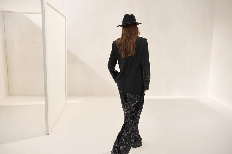 Back view of a fashionable woman wearing a black blazer, sequined pants, and a fedora hat in a bright white environment.