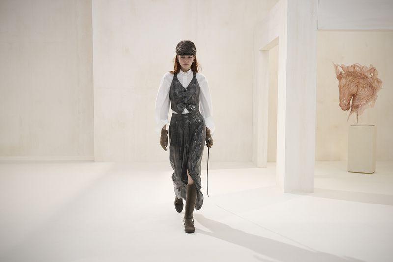 A woman in a vintage-inspired equestrian outfit with a riding crop walks in a bright white interior, showcasing fashion-forward style.