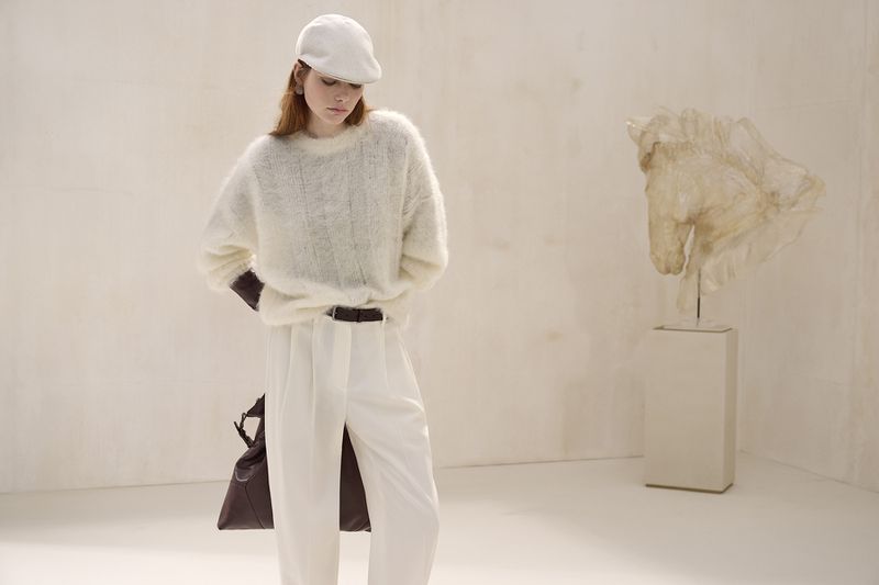 A young woman stands in a studio with a horse sculpture, modeling a white fuzzy sweater, white pants, cap, and a brown leather shoulder bag.