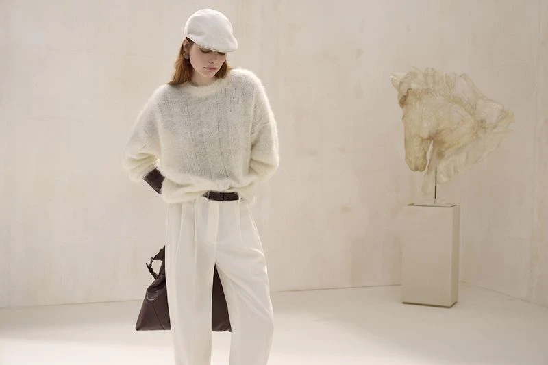 A young woman stands in a studio with a horse sculpture, modeling a white fuzzy sweater, white pants, cap, and a brown leather shoulder bag.