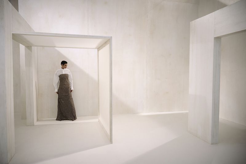Elegant woman posing in a long gown with white shirt in a minimalistic white interior space, creating a surreal, fashion-forward image.
