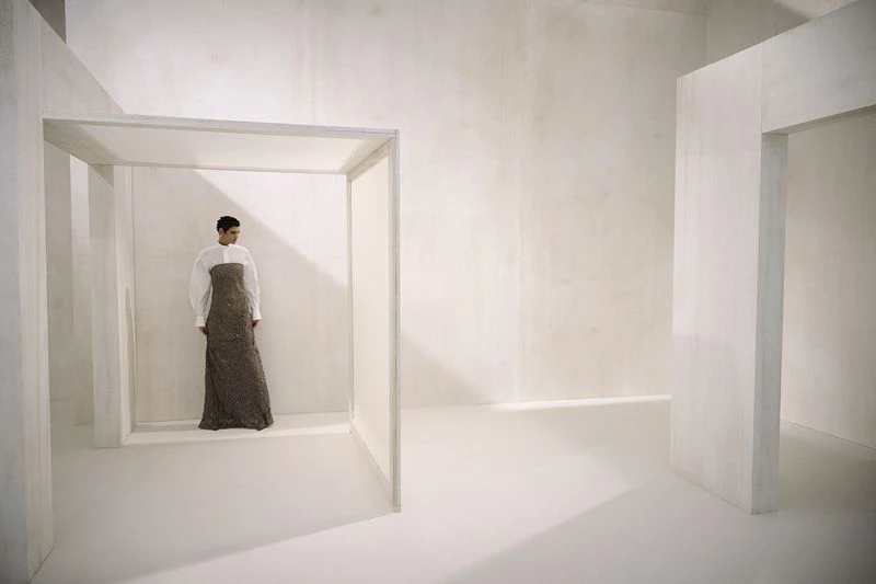 Elegant woman posing in a long gown with white shirt in a minimalistic white interior space, creating a surreal, fashion-forward image.