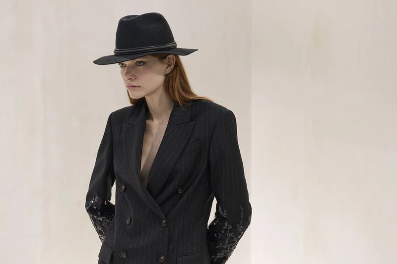 Adult woman in pinstripe blazer and fedora hat poses in a fashion shot in a studio with a neutral color wall in the background.