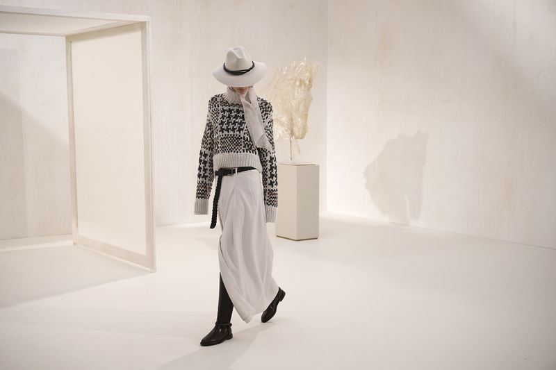 Stylish woman in a patterned sweater, white skirt, and hat walks in a bright studio with an abstract sculpture in the background.