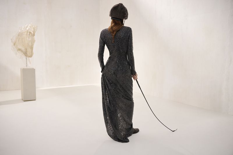 An adult woman in a long, dark, sequined dress and riding hat holds a riding crop in a light-filled room with an abstract sculpture.