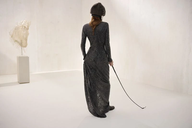 An adult woman in a long, dark, sequined dress and riding hat holds a riding crop in a light-filled room with an abstract sculpture.
