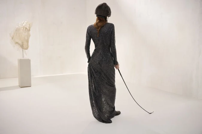 An adult woman in a long, dark, sequined dress and riding hat holds a riding crop in a light-filled room with an abstract sculpture.