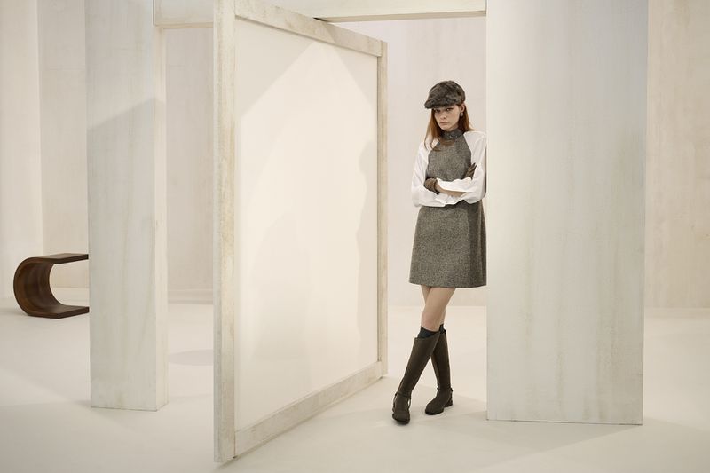 A fashionable woman in a gray dress and hat poses in a bright white room with architectural details, complemented by brown knee-high boots.