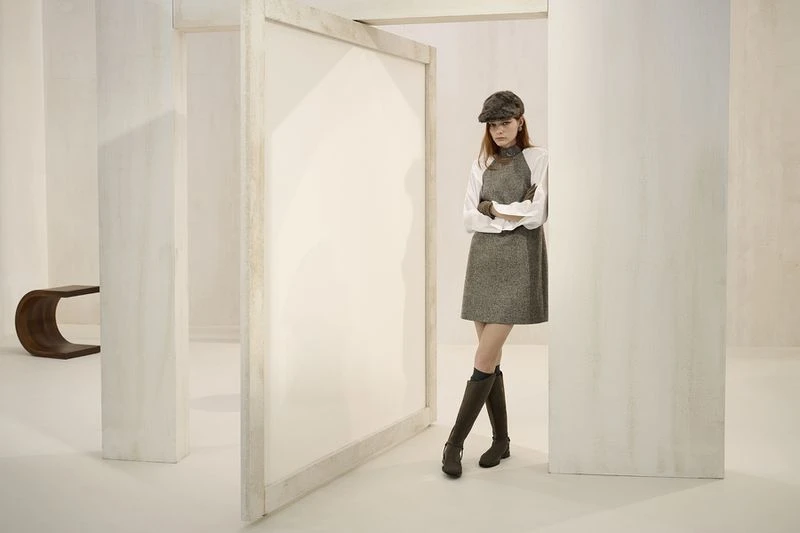 A fashionable woman in a gray dress and hat poses in a bright white room with architectural details, complemented by brown knee-high boots.