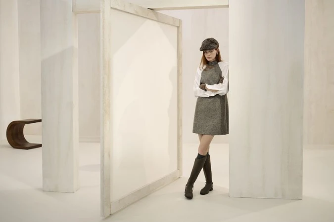 A fashionable woman in a gray dress and hat poses in a bright white room with architectural details, complemented by brown knee-high boots.