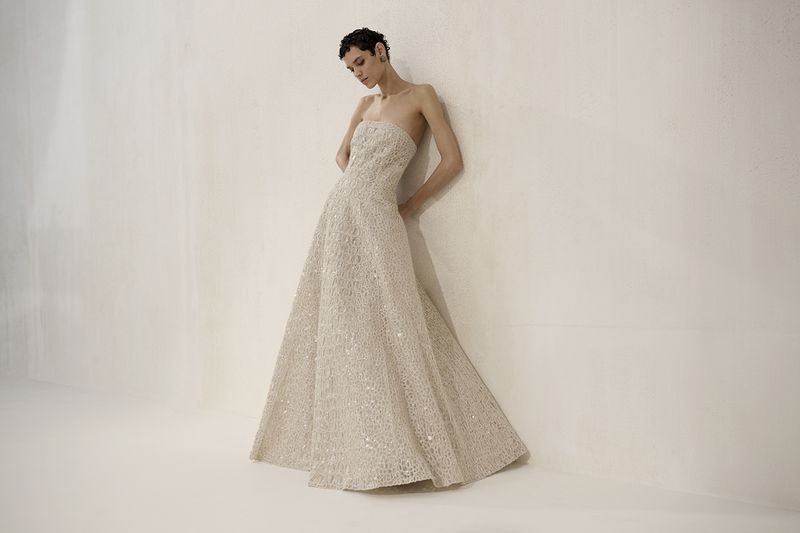 Elegant adult female in a strapless formal gown with a full skirt, leaning against a wall in a neutral-toned studio setting.