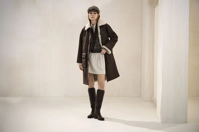 Full-length studio shot of a woman in a dark brown shearling coat, cream skirt, and knee-high boots against an off-white backdrop.