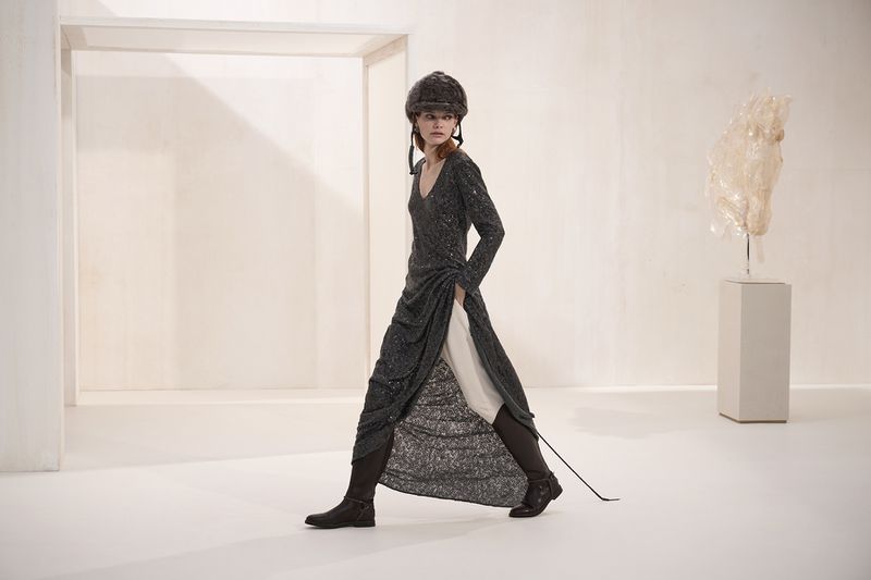 A woman in a grey dress and furry hat walks in a bright white space, with a sculpture on a pedestal in the background, fashion editorial.