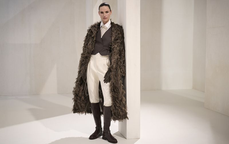 A fashionable adult female poses in a photo studio. She wears a long brown faux fur coat, vest, gloves, and white breeches with tall brown boots.
