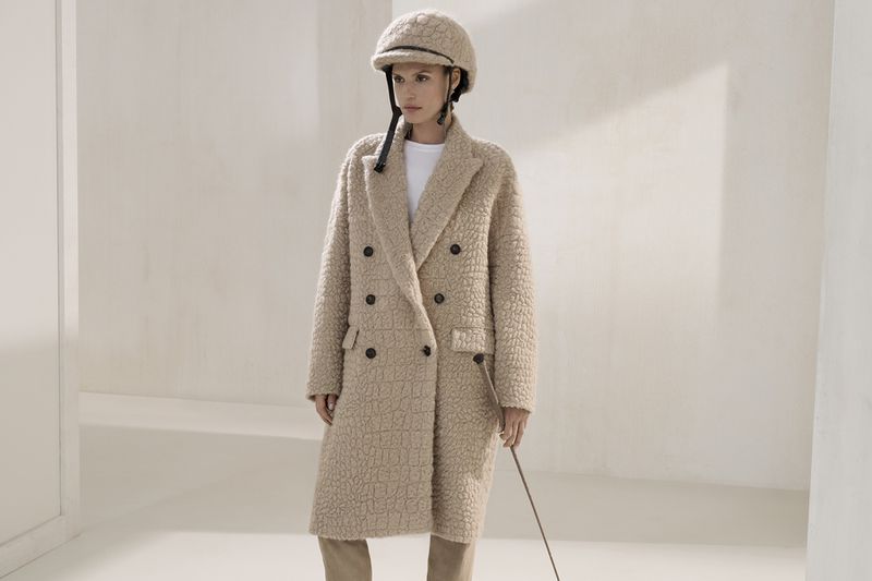 A woman poses in a double-breasted coat with a riding helmet, standing in an off-white indoor setting. The light is soft and diffused.