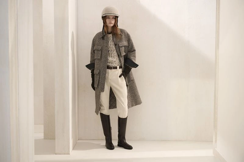 A woman stands indoors wearing riding boots, cream-colored riding pants, a tweed overcoat, gloves and a riding helmet against a white wall.
