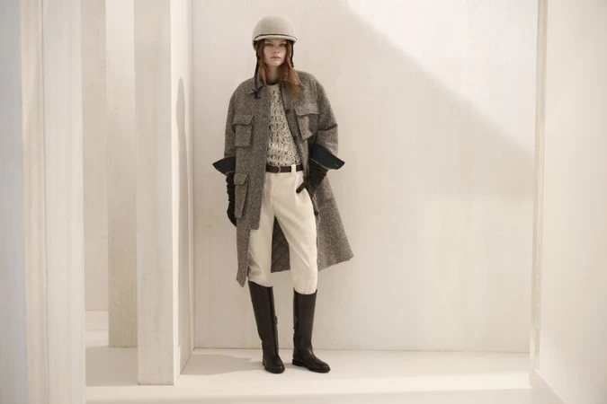 A woman stands indoors wearing riding boots, cream-colored riding pants, a tweed overcoat, gloves and a riding helmet against a white wall.