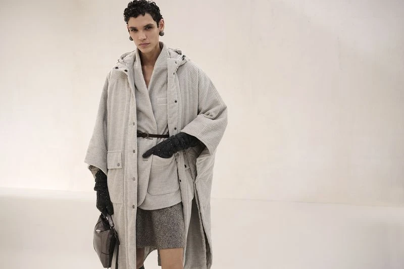 Fashionable woman in grey coat and skirt holding purse in studio, stylish contemporary outfit. Cool-toned lighting, elegant.