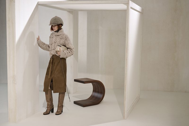 Fashion shoot featuring an adult female wearing a beige knit sweater, brown skirt, hat, and boots in a bright, minimalist indoor space.