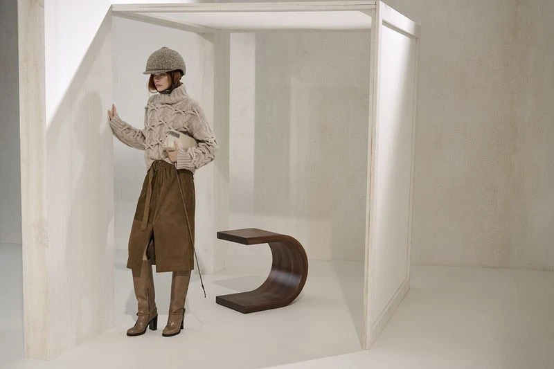 Fashion shoot featuring an adult female wearing a beige knit sweater, brown skirt, hat, and boots in a bright, minimalist indoor space.
