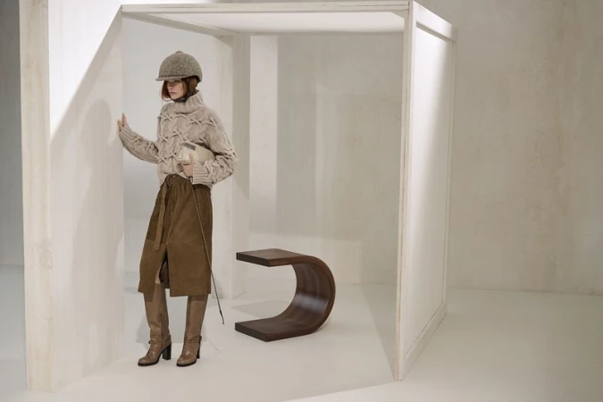Fashion shoot featuring an adult female wearing a beige knit sweater, brown skirt, hat, and boots in a bright, minimalist indoor space.