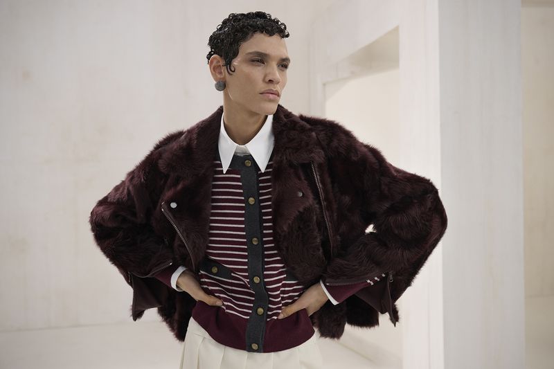 Fashionable adult with dark, curly hair wearing a striped sweater, white collared shirt, and faux fur coat poses indoors with hands on hips.