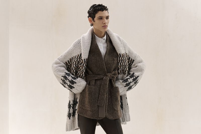 Female model wearing a brown belted jacket and patterned knit cardigan in a fashion editorial studio shot.