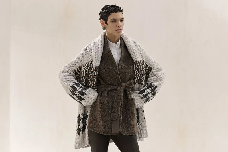 Female model wearing a brown belted jacket and patterned knit cardigan in a fashion editorial studio shot.