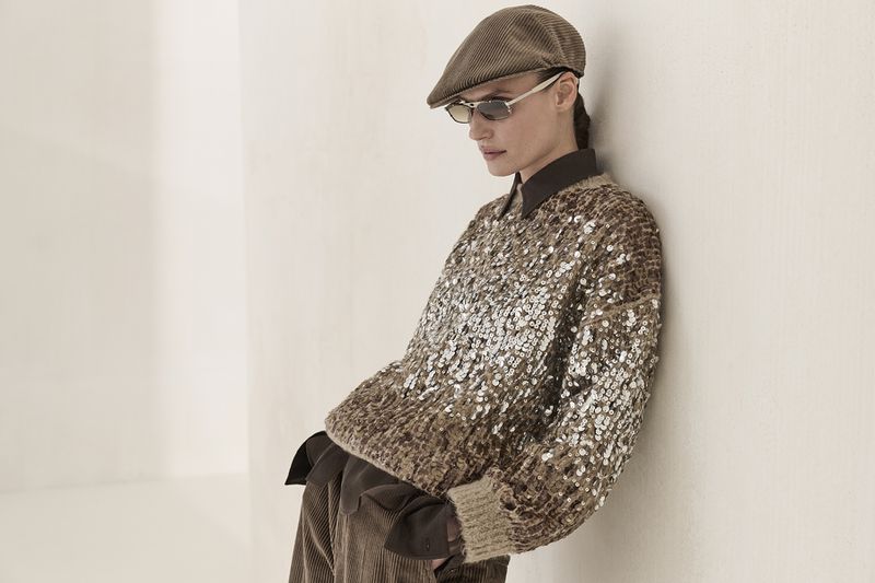 A woman in a brown sequined sweater, corduroy pants and cap leans against a white wall in a fashion shot with soft, neutral lighting.