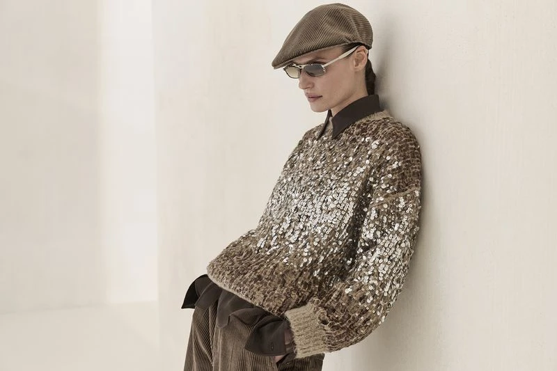 A woman in a brown sequined sweater, corduroy pants and cap leans against a white wall in a fashion shot with soft, neutral lighting.