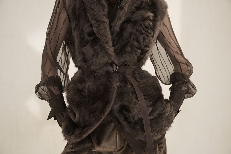Fashionable woman wearing a brown fur vest, corduroy skirt, and striped sheer sleeves posing against a neutral background.