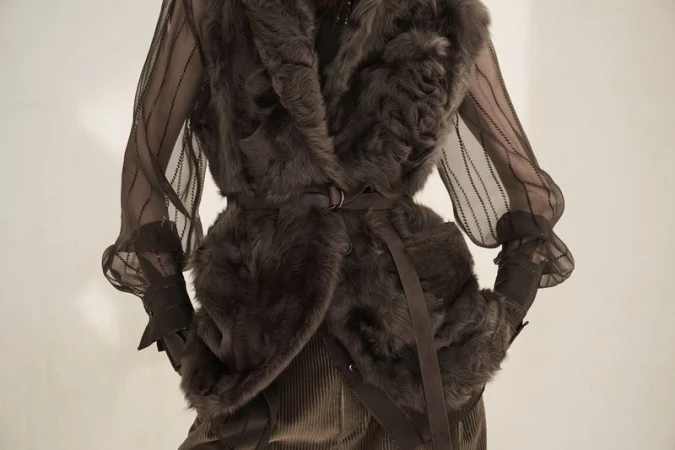 Fashionable woman wearing a brown fur vest, corduroy skirt, and striped sheer sleeves posing against a neutral background.