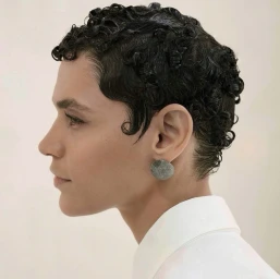Profile portrait of a model with short curly hair wearing a white collared shirt and textured round earring against a neutral backdrop.